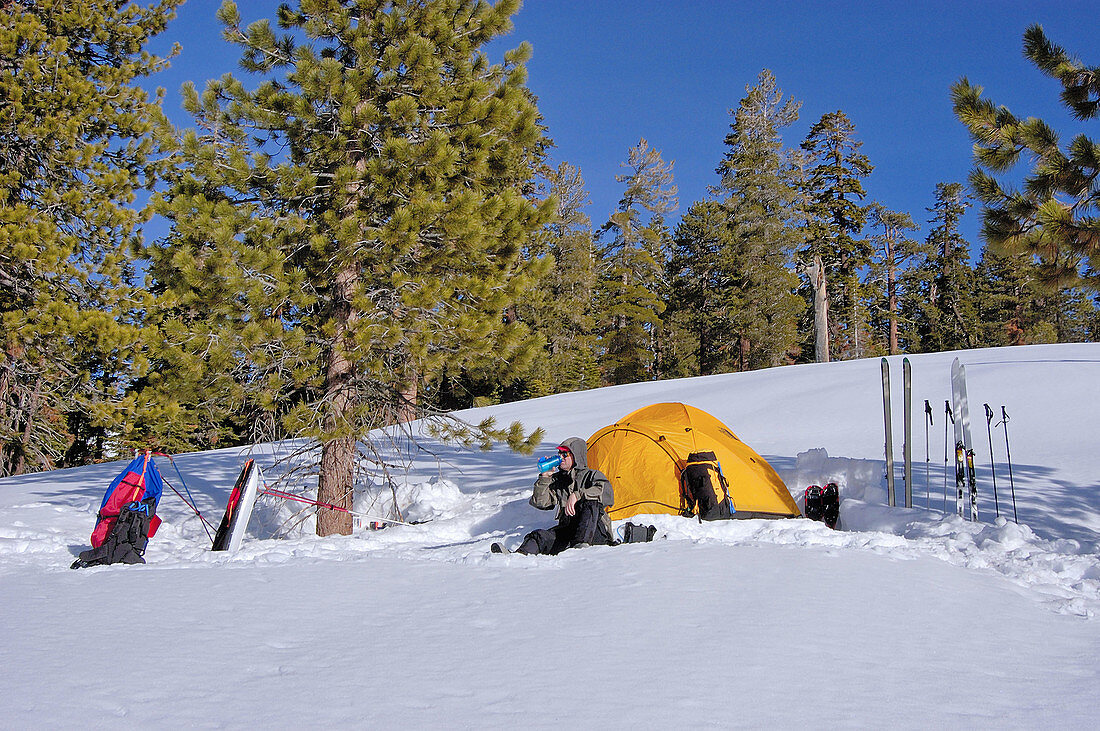 Skier drinking from a bottle in front of a yellow dome tent in a backcountry ski camp, Yosemite National Park, California