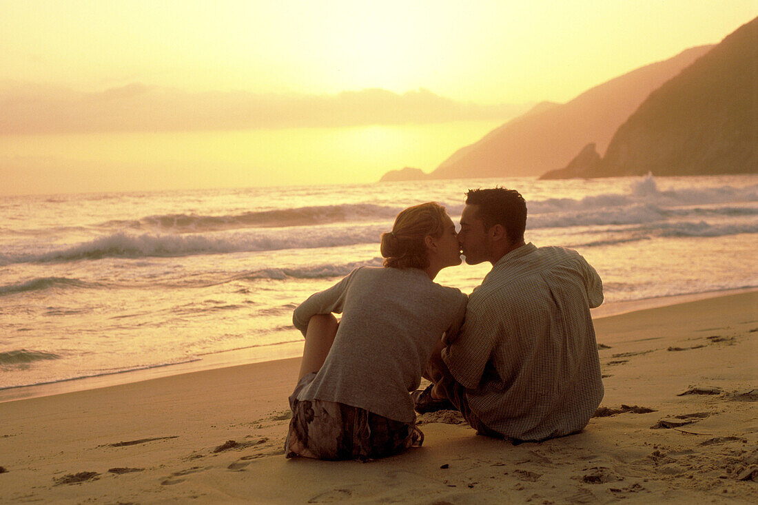 Young couple sitting on sand at the beach kissing at sun set