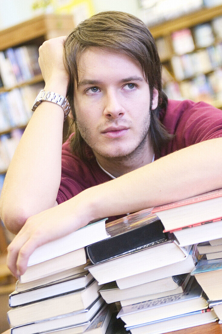 24 year old man leaning over a pile of books, head in hand, in the library