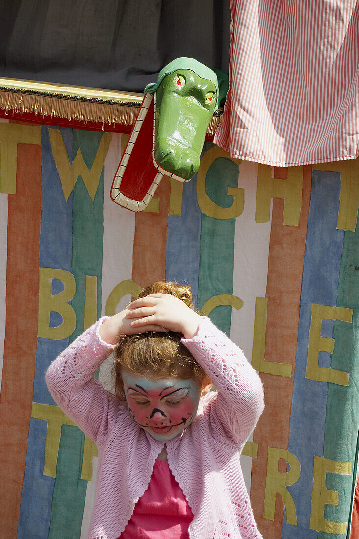 3 year old girl standing in front of a Punch and Judy stall, with a crocodile snapping at her head.