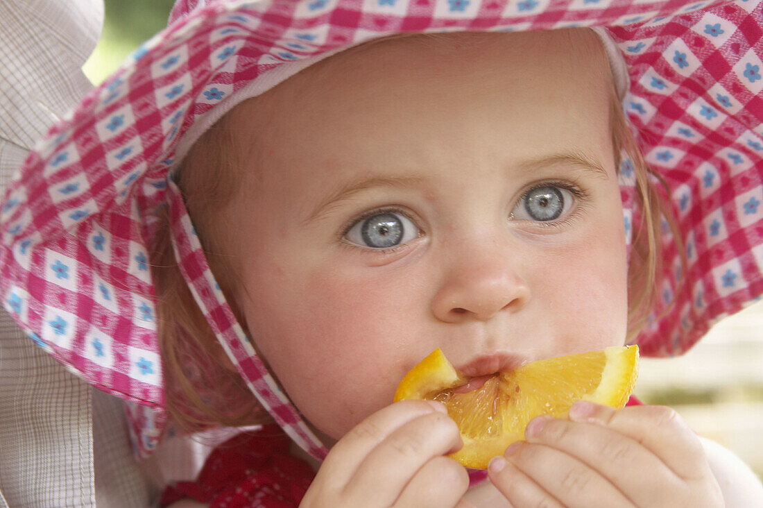 Headshot of an 18 month old girl wearing a big floppy sunhat, eating a slice of orange.