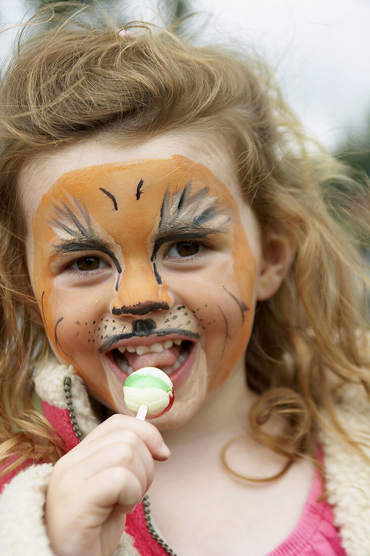 3 year old girl with her face painted, looking at the camera, smiling, eating a lolly