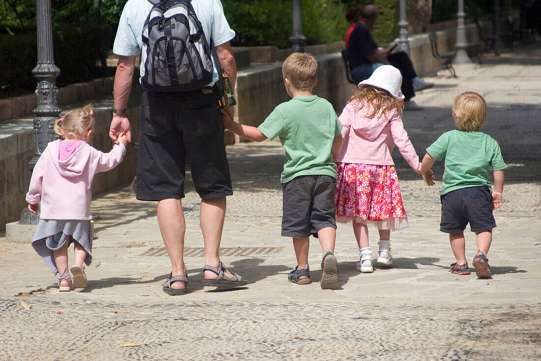The back and legs of a man walking down a street holding four small childrens hands, all walking in a row