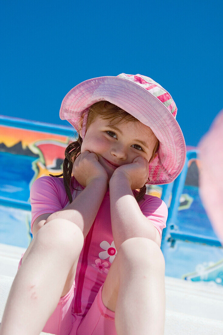 5 year old girl, sitting smiling into camera, wearing a swimsuit and sunhat, at the swimming baths