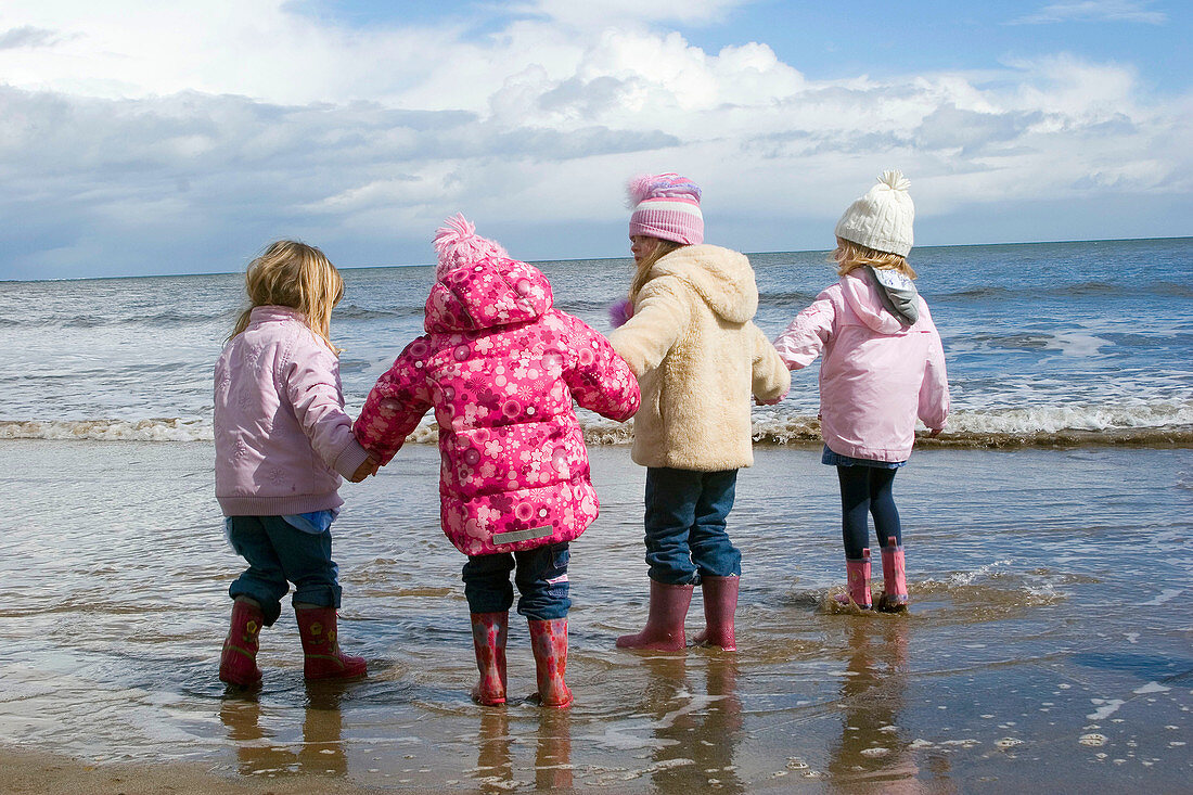 Group of 4, 4 year old girls standing on the seashore all in a row holding hands, dressed in winter clothing.