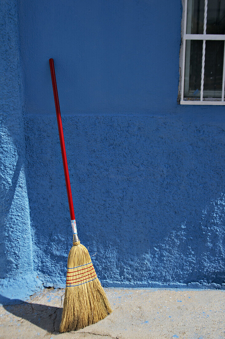 Straw broom with red handle lean against blue stucco exterior wall, metal frame over window. La Paz. Mexico.