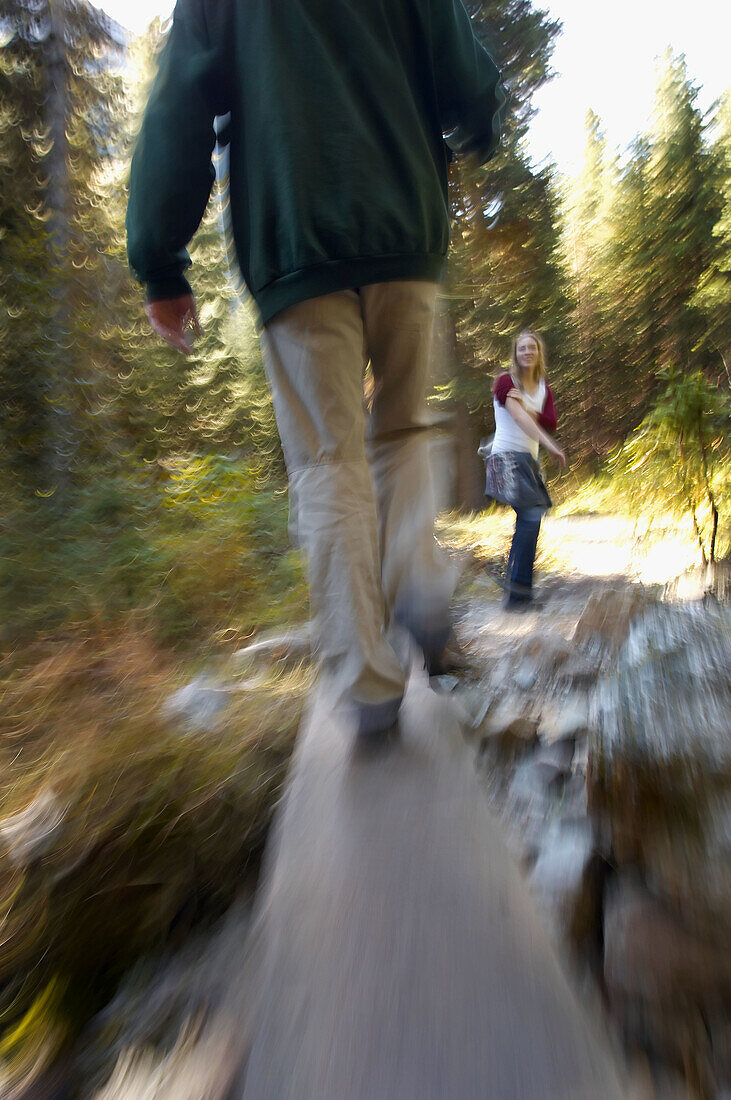 couple hiking on mountain trail with man walking on a log across a stream
