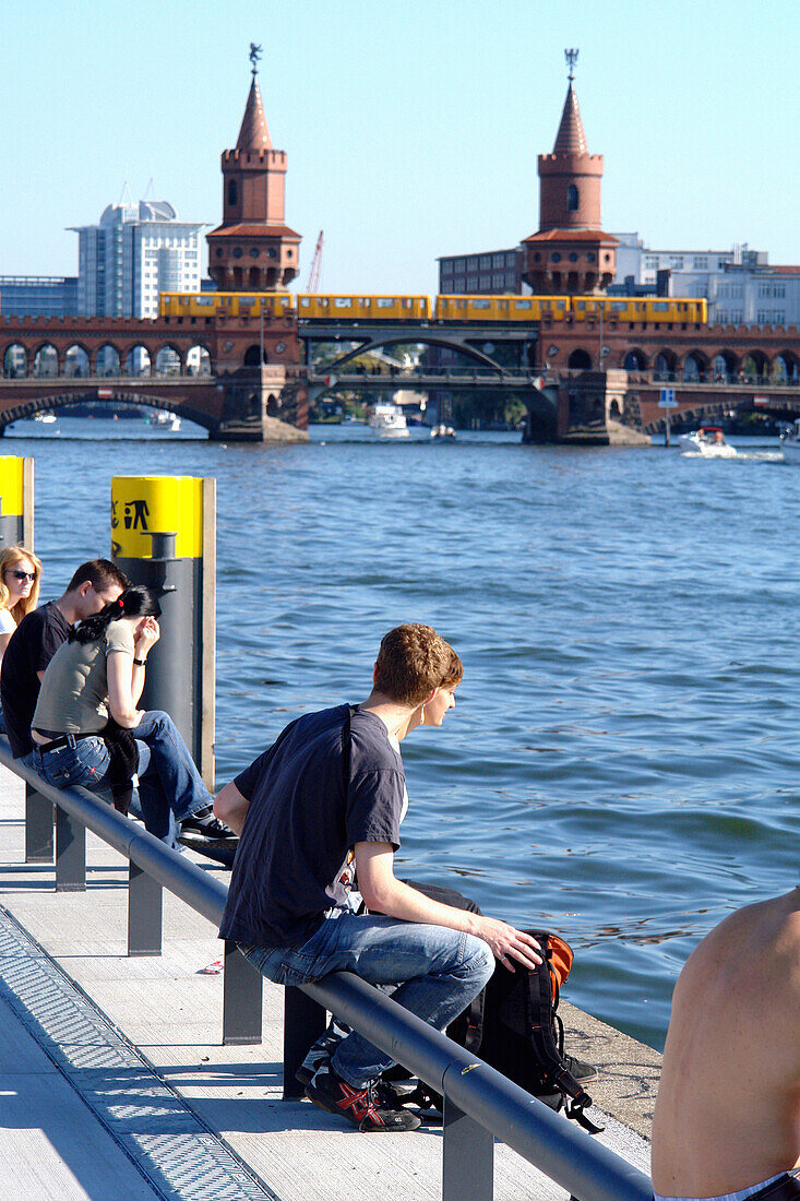 Young people sitting at Spree riverside, Oberbaum Bridge in background, Berlin, Germany