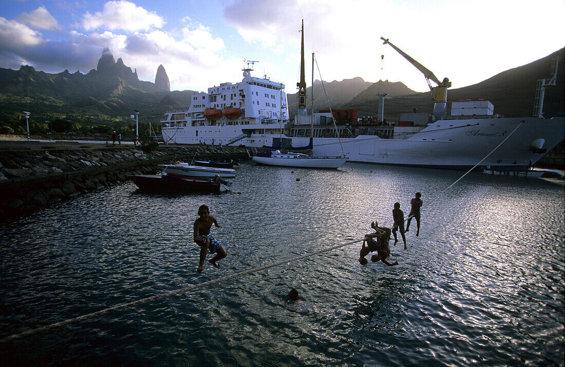 The Aranui III anchoring in the harbour of Hakahau on the island of Ua Pou, children jumping from arope into the water, French Polynesia