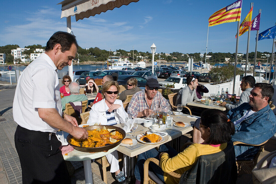 Kellner serviert Paella auf Terrasse des La Caracola Restaurant, Portocolom, Mallorca, Balearen, Spanien, Europa