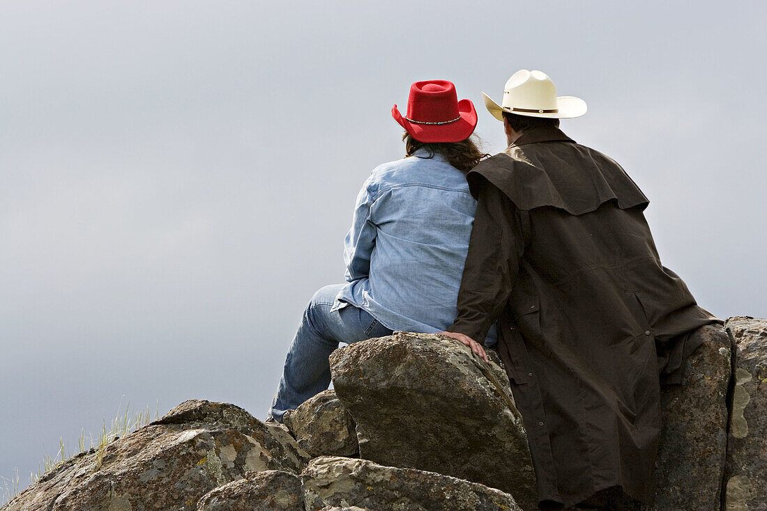 Female and male in western attire sitting together on the rocks out in the country looking off into the distance beyond them.