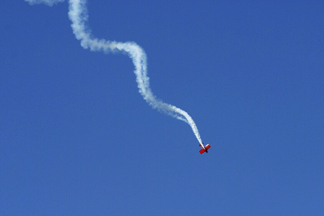 Bright red stunt plane flying downward in a spiral against a clear blue sky at an air show