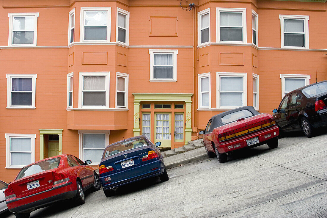 Cars parked in front of a bright, well-maintained orange house on a very steeply sloped section of Jones Street in San Francisco, California. The street is so steep that the sidewalk has steps.