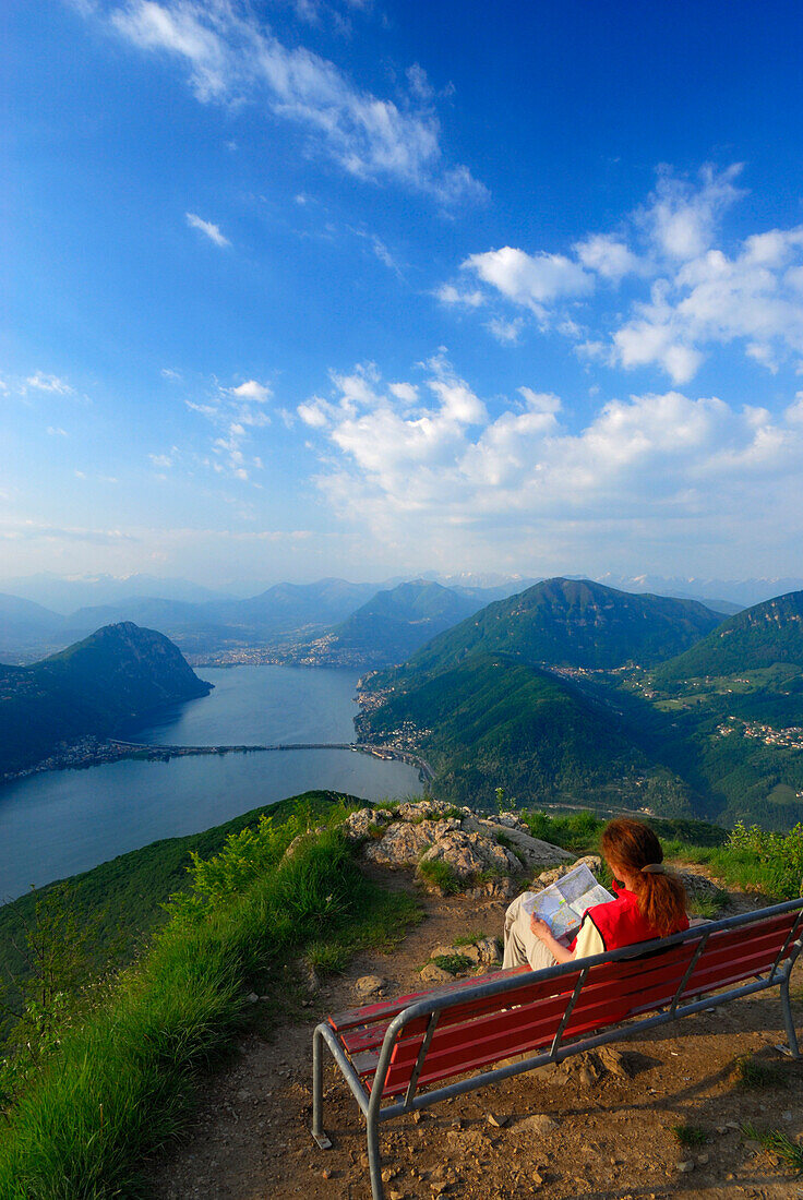 Mid adult woman sitting on a bench, Lake Lugano in background, Monte San Giorgio, Ticino, Switzerland
