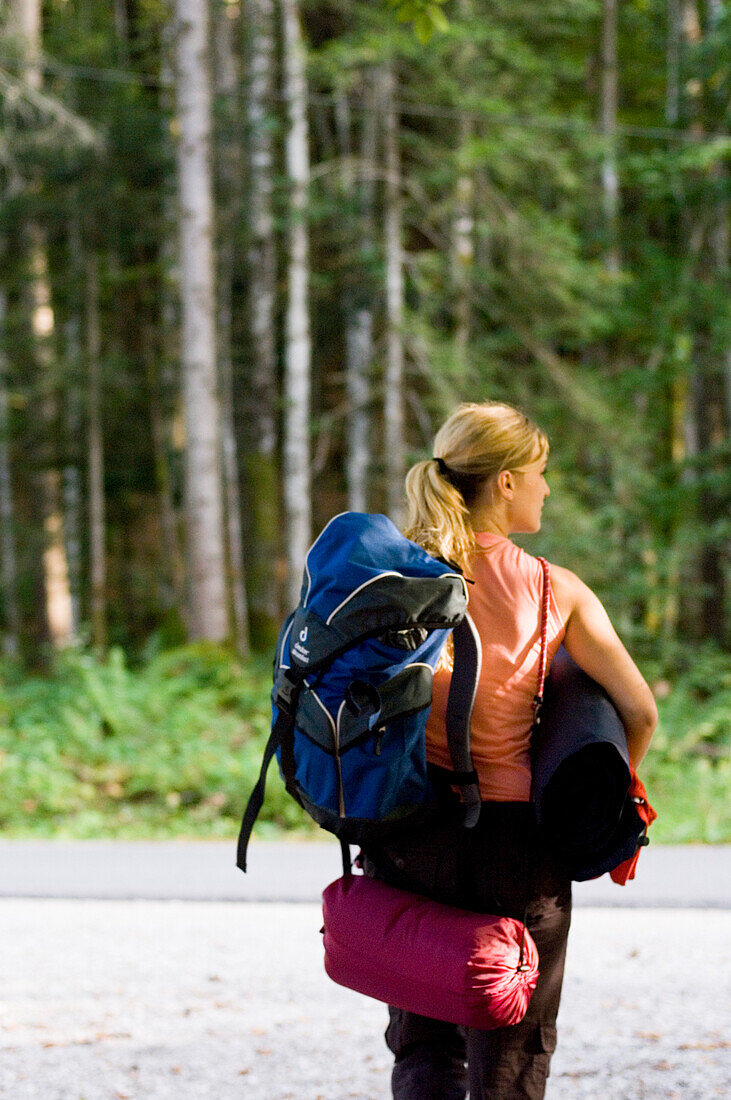 Junge Frau mit Rucksack, Schlafsack und Isomatte wandert durch einen Wald, Bayern, Deutschland