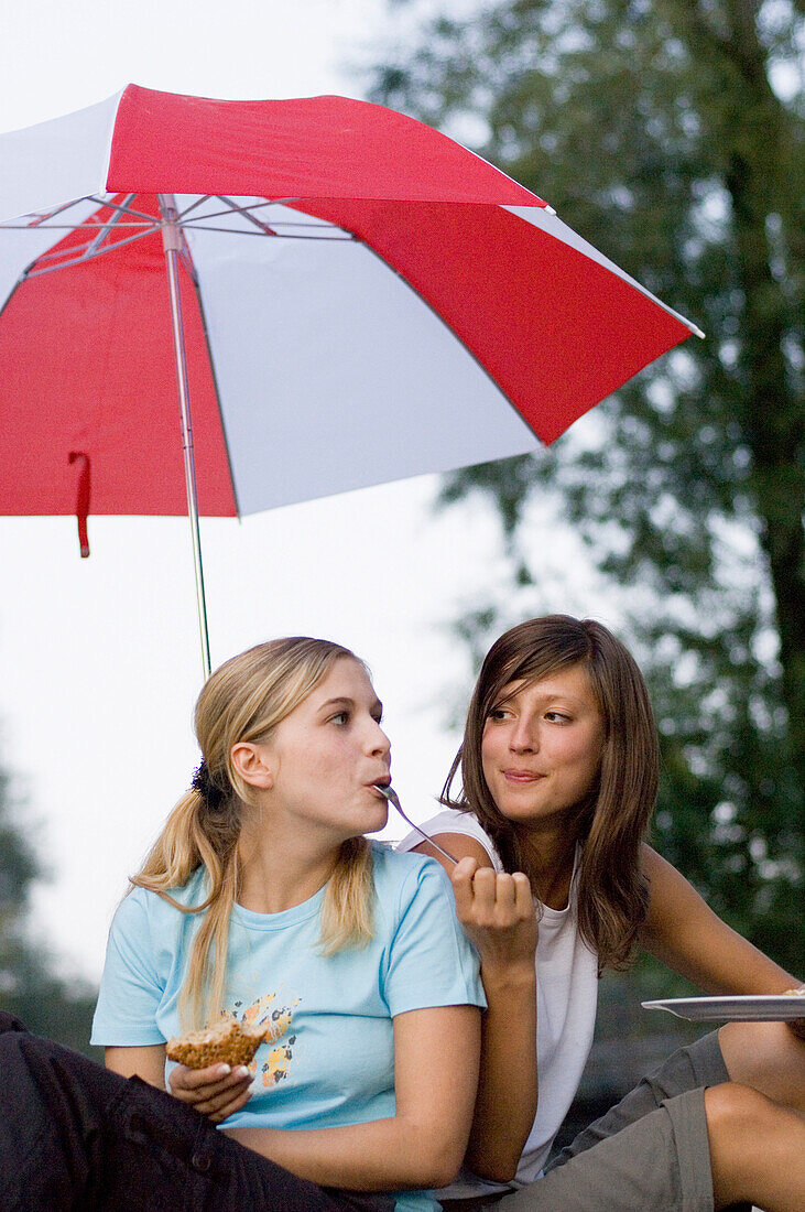 Two young women sitting under an umbrella while eating, Munich, Bavaria, Germany
