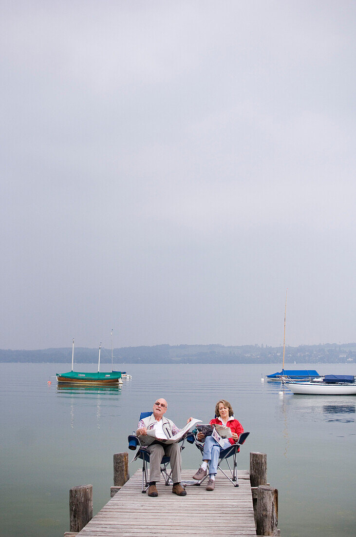 Older couple sitting on the jetty reading a newspaper, Lake Ammersee, Bavaria, Germany