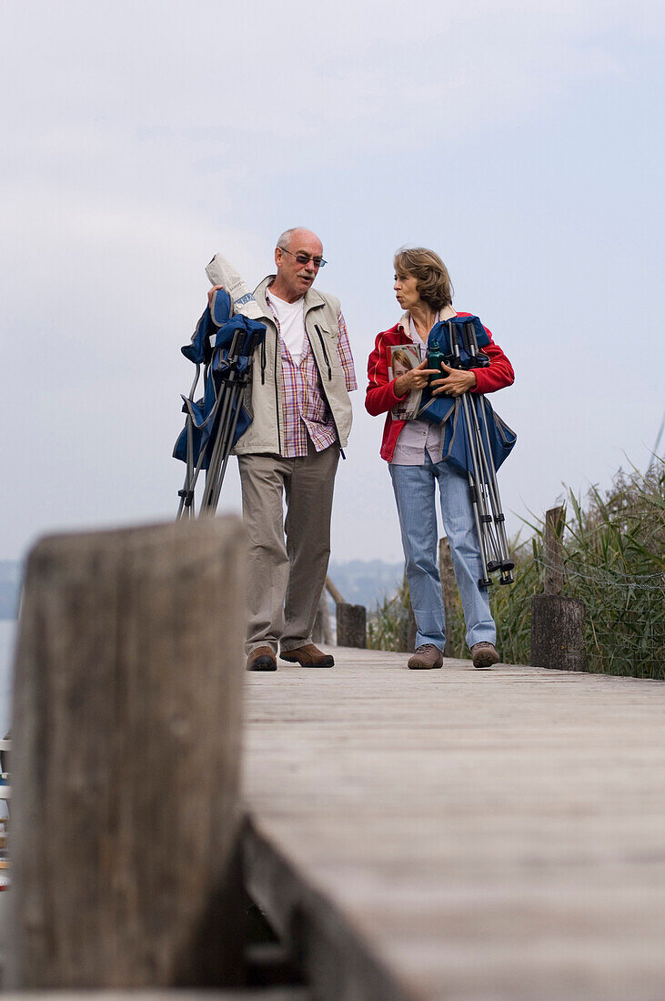 Senior couple carrying folding chairs along a jetty, lake Ammersee, Bavaria, Germany
