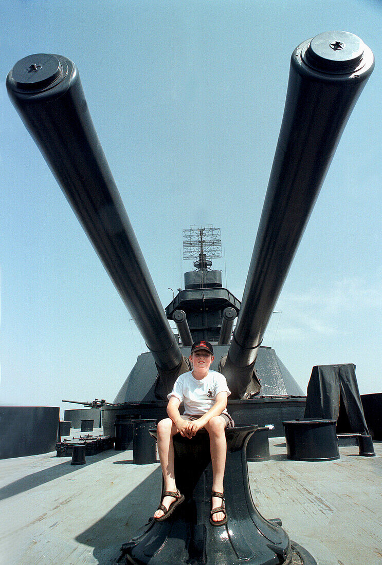 A boy visits the WWII USS Texas Battleship on display near Houston Texas.  He sits between some deck guns for a photograph.