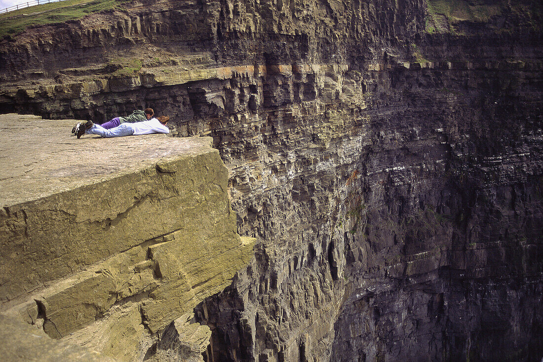 Two people lie precariously on the edge of great slab overlooking the Cliffs of Moher near Lahinch. Co. Clare, Ireland