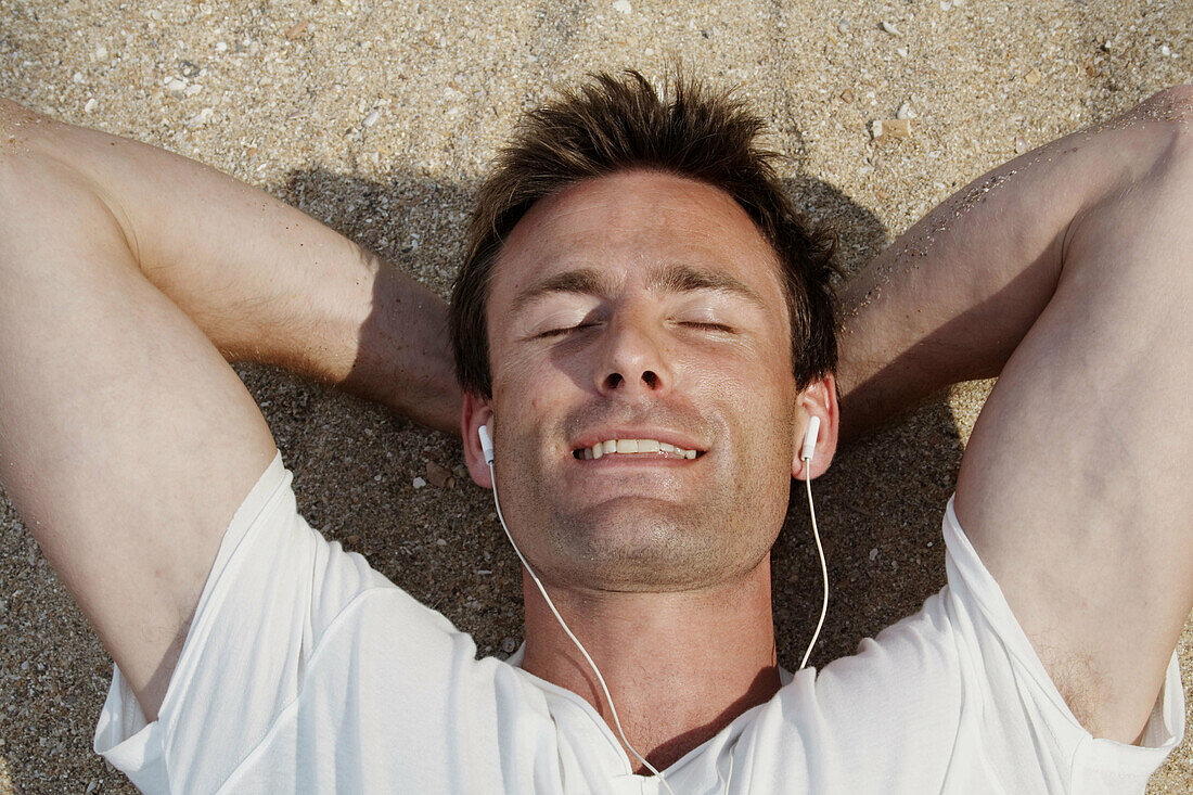 Horizontal shot of a god-looking man lying on the beach in India and listening to headphones with his eyes closed.