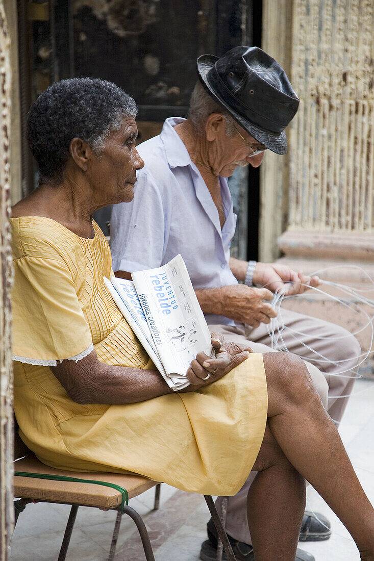 Couple of old people sitting in front of their home (Habana Vieja). Selling the newspaper Juventud revelde. Havana. Cuba.