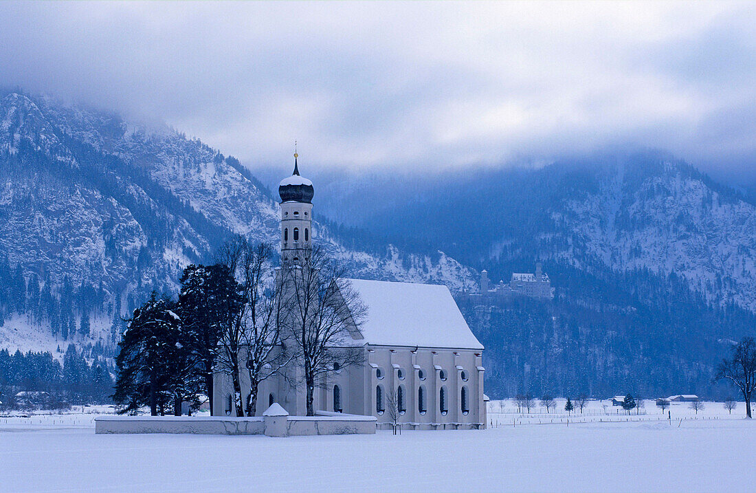 Europe, Germany, Bavaria, Schwangau near Füssen, St. Coloman pilgrimage church surrounded by trees and Neuschwanstein Castle in the mountains