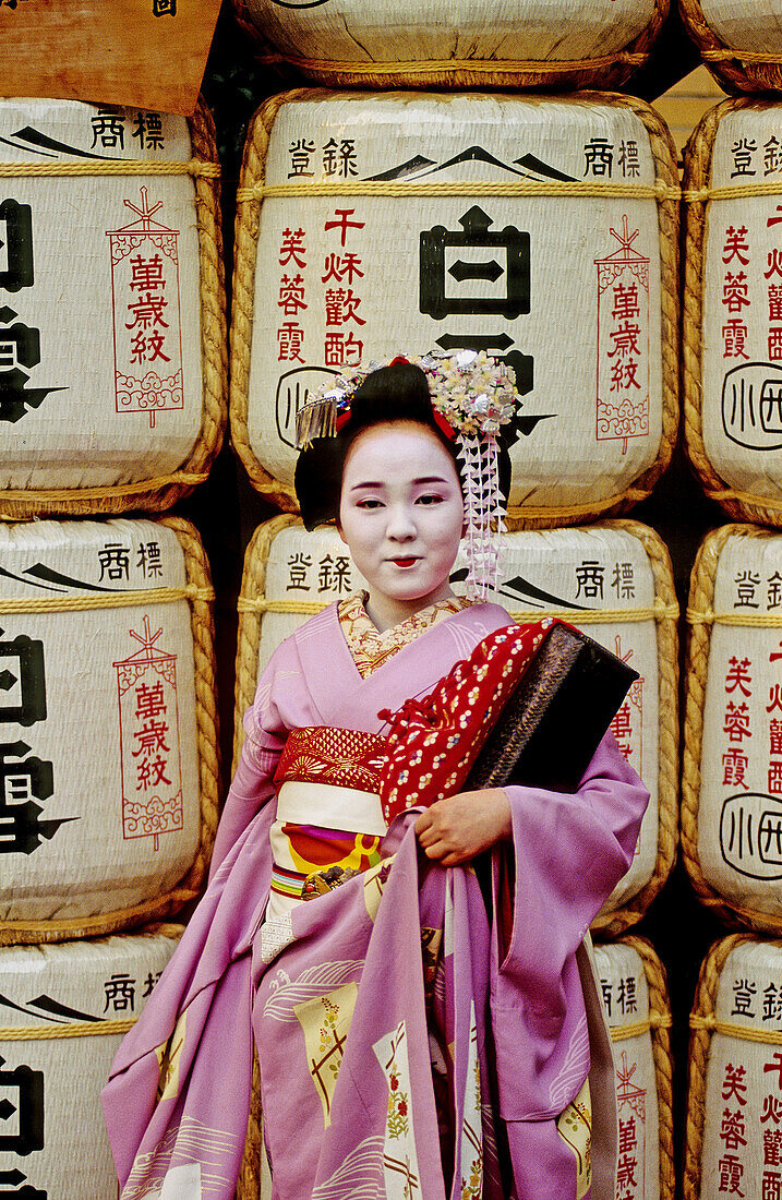 16 year old 'maiko' (geisha apprentice) standing by sake barrels in a Gion shinto temple. Kyoto, Japan