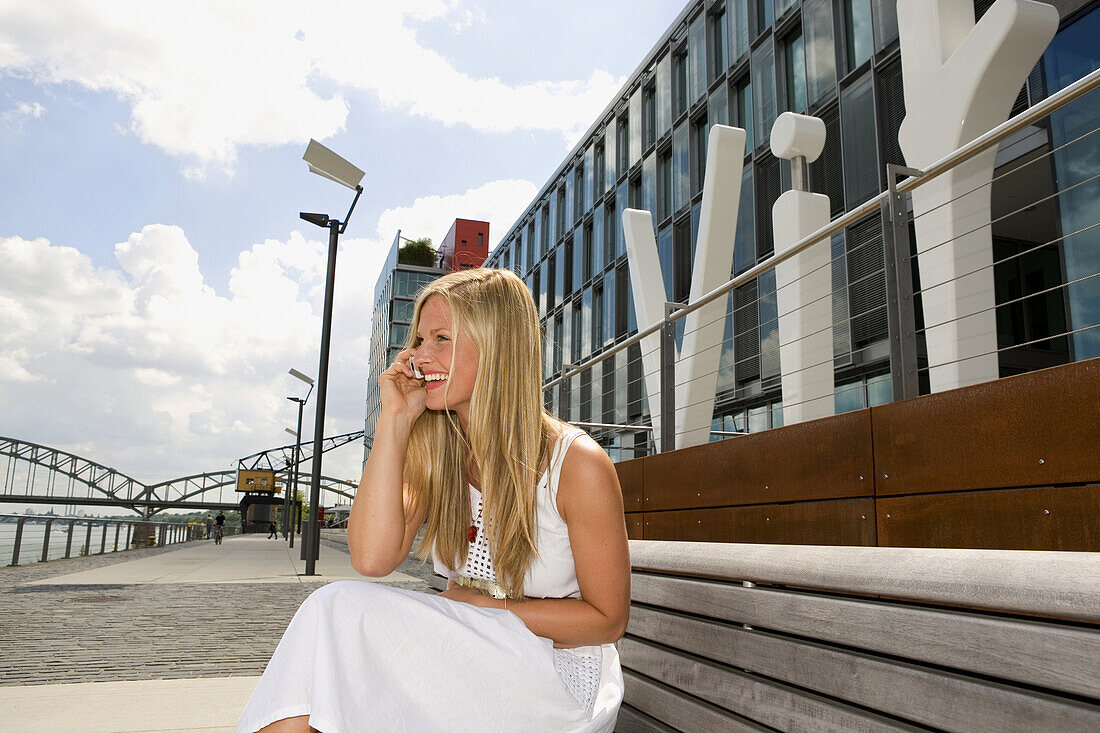 Junge Frau sitzt auf Bank und telefoniert, Köln, Nordrhein-Westfalen, Deutschland