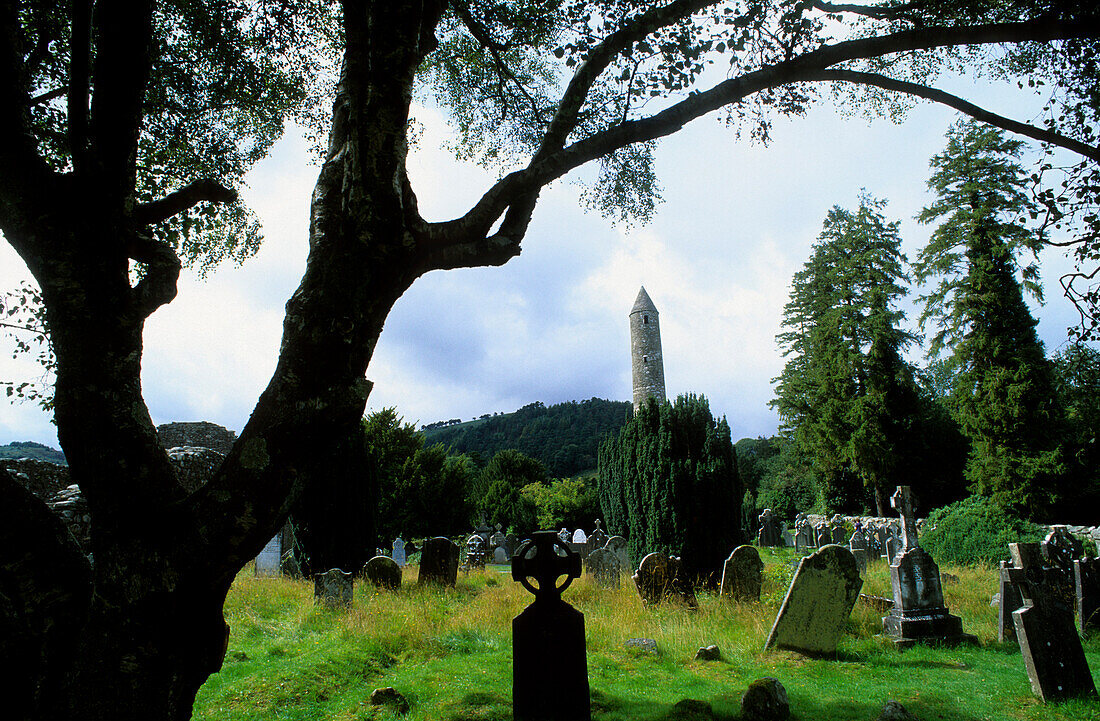 Round bastion and weathered graveyard under trees, Glendalough, County Wicklow, Ireland, Europe