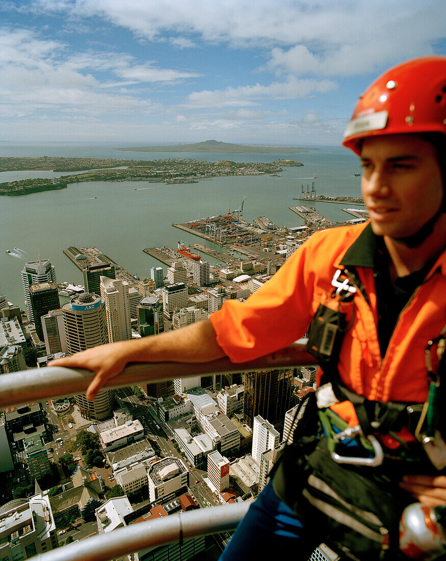 Junger Mann mit Kletterausrüstung auf dem Sky Tower, Blick auf Central Business District und Waitemata Harbour, Auckland, Nordinsel, Neuseeland