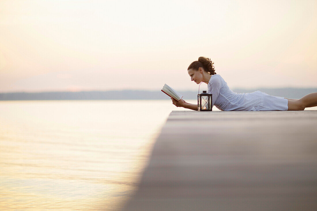 Frau liegt auf einem Steg am Starnberger See und liest ein Buch, Ambach, Bayern, Deutschland