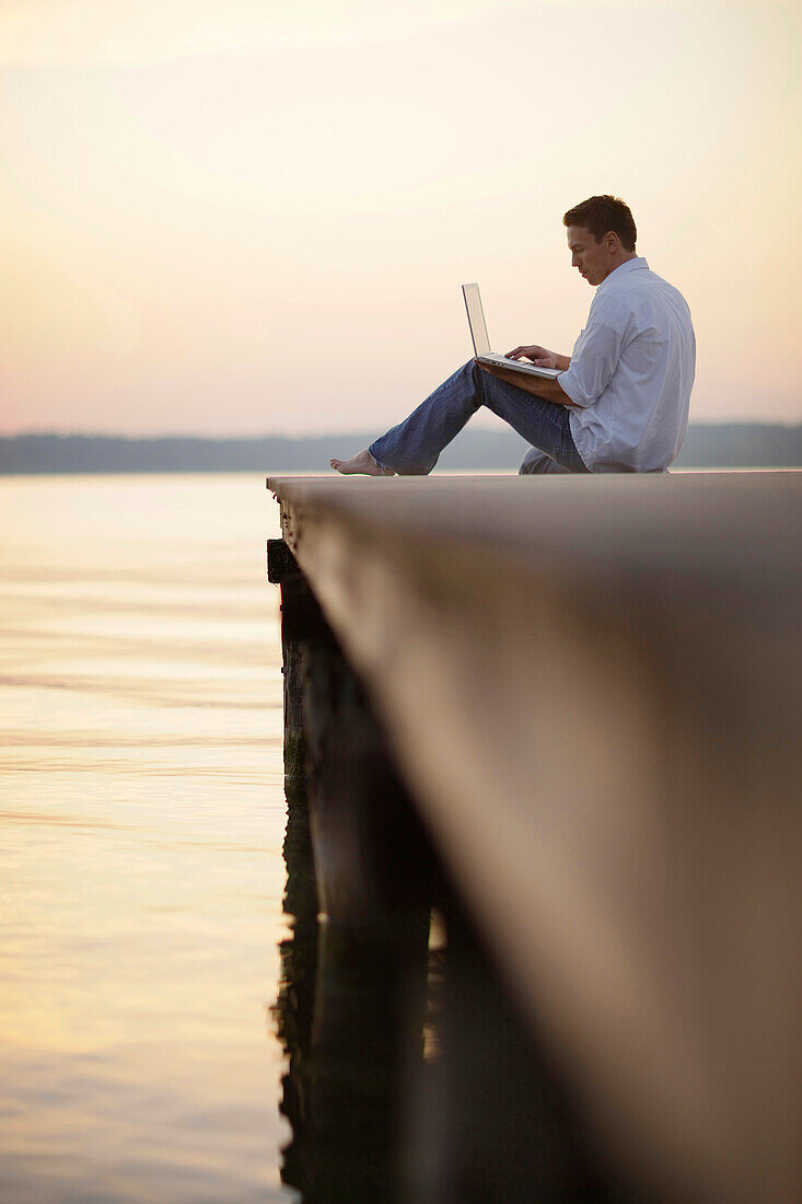 Mann mit Laptop sitzt auf einem Steg am Starnberger See, Ambach, Bayern, Deutschland