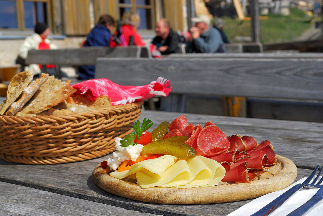 South Tyrolean snack with South Tyrolean speck, bacon, salami, peperoni, pepperoni, cheese and Schuettelbrot, group of people out of focus in background on terrace of hut Radlseehuette, Sarntaler Alpen, Sarntal range, South Tyrol, Italy