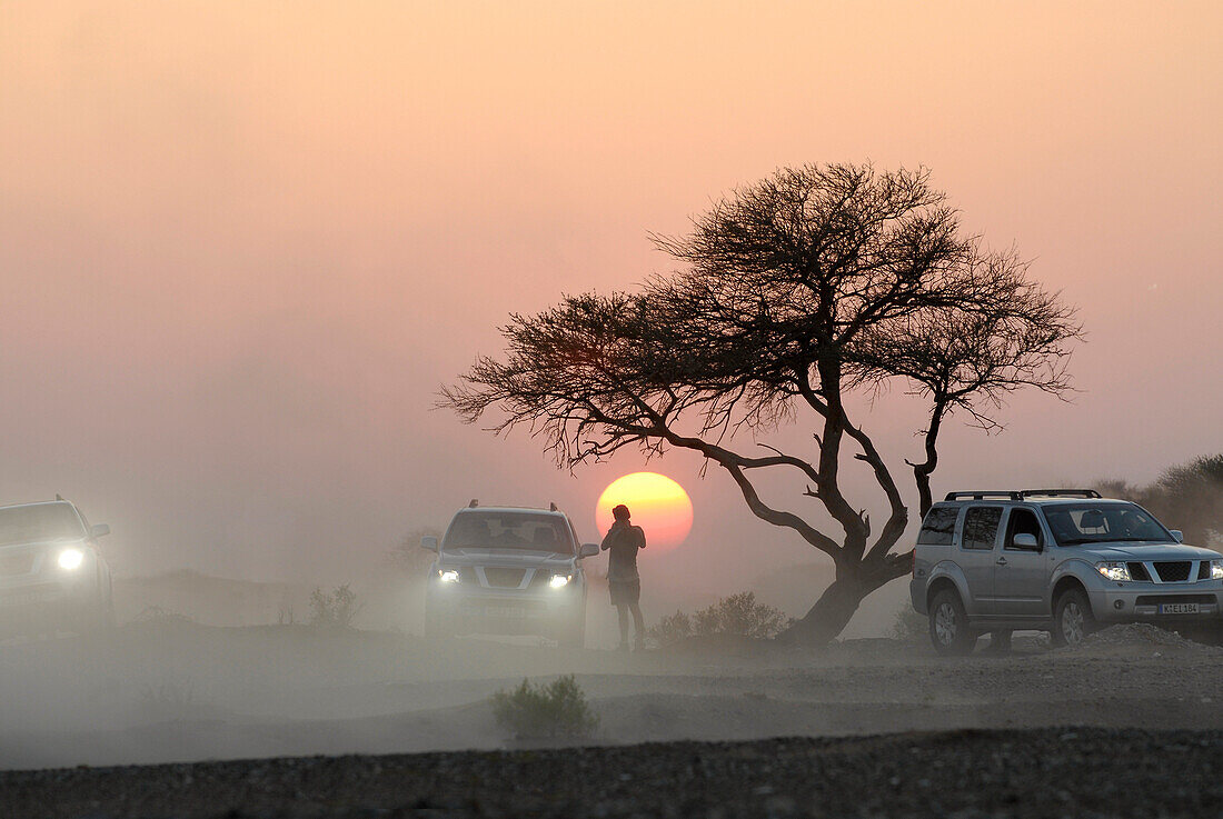 Eine Person und drei Geländewagen vor der untergehenden Sonne, Al Hajar Berge, Oman, Asien