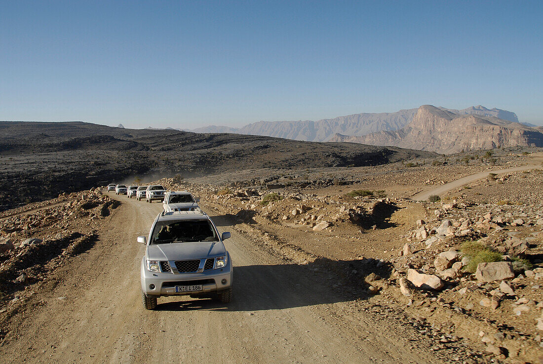 A row of all-terrain vehicles driving on a road through barren scenery, Al Hajar mountains, Oman, Asia