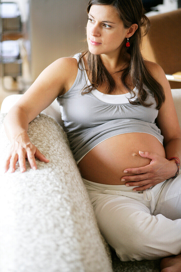 Schwangere Frau sitzt einer Couch und berührt ihren Bauch