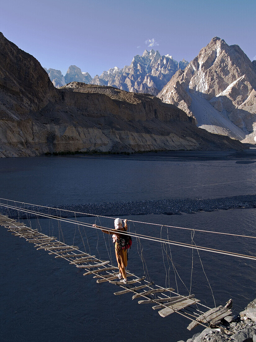 Trekker makes the crossing of the Hunza River in the Karakoram Mountains of Pakistan by rickety footbridge