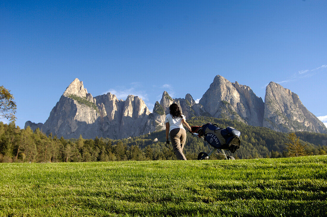 Frau mit Golftasche auf dem Rasen unter blauem Himmel, Golfplatz Kastelruth Seiser Alm, Schlern, Südtirol, Italien, Europa