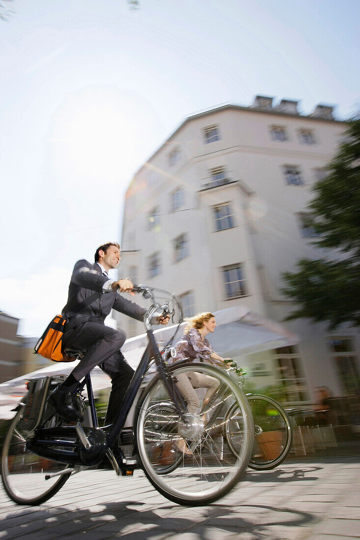 Two businesspeople riding bicycles over cobblestone pavement, Munich, Bavaria, Germany