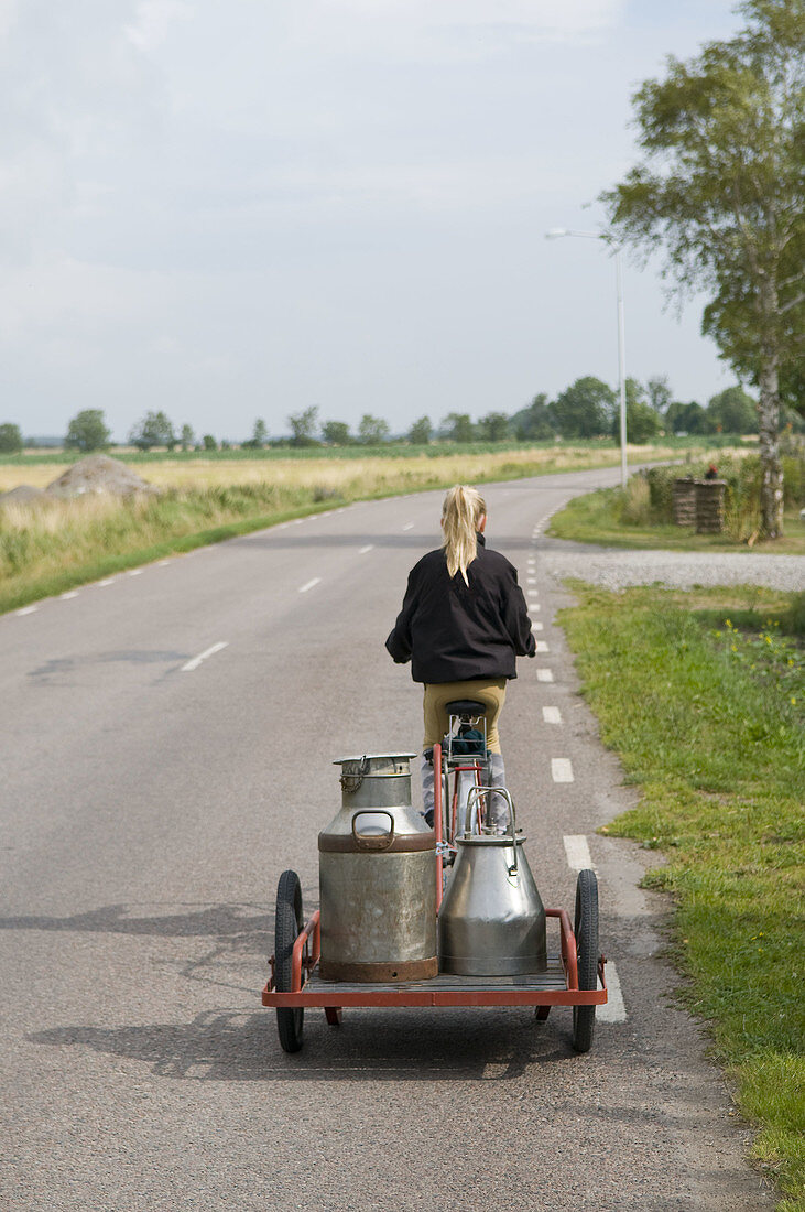 Girl on a bicycle dragging a cart with milkbottles.