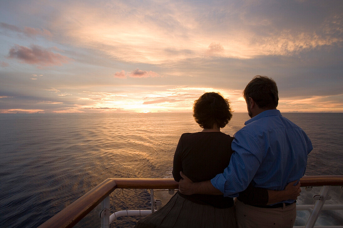 Couple standing at the railing of MV Columbus looking at sunset, South Pacific, Oceania