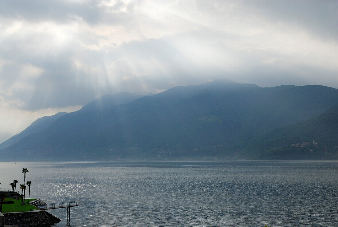Lichtstrahlen durch Wolken über Lago Maggiore, Strandbad mit Palmen, Brissago, Lago Maggiore, Tessin, Schweiz