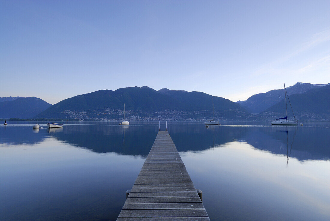 Holzsteg führt in Lago Maggiore mit ankernden Segelbooten mit Locarno, Muralto und Minusio am jenseitigen Ufer, Gambarogno, Lago Maggiore, Tessin, Schweiz