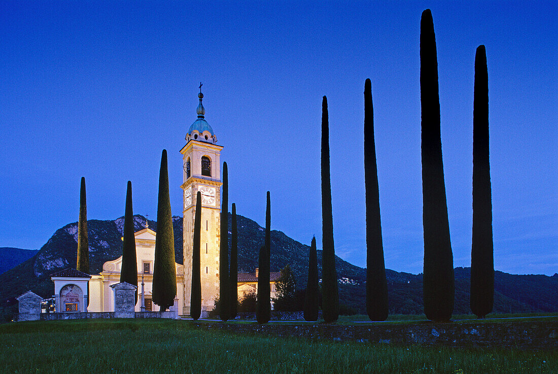 Alley with cypresses to the illuminated church Sant´Abbondio in the evening, Ticino, Switzerland, Europe