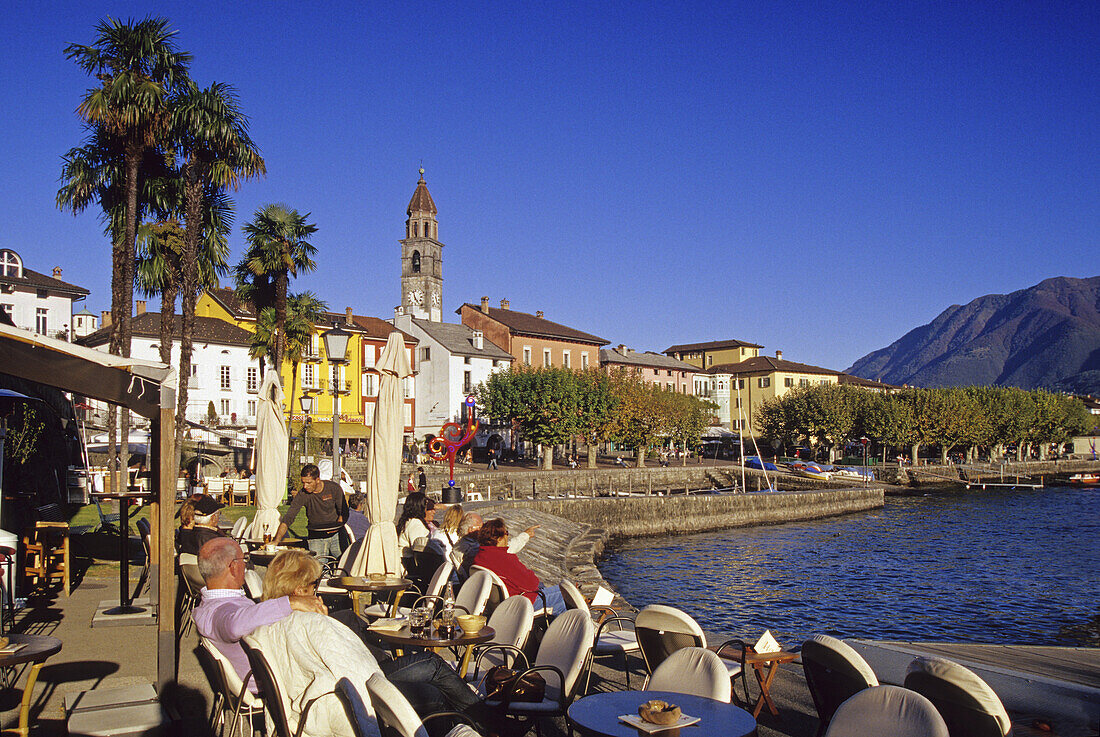 Menschen in einem Café am Hafen unter blauem Himmel, Ascona, Lago Maggiore, Tessin, Schweiz, Europa