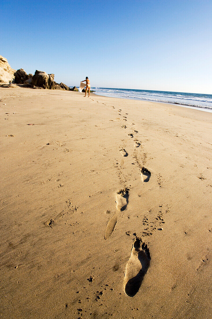 Fußspuren am Strand mit einer schwangeren Frau im Hintergrund die ihr Handtuch auslegt, Strand von Conejo, Baja California süd, Mexiko