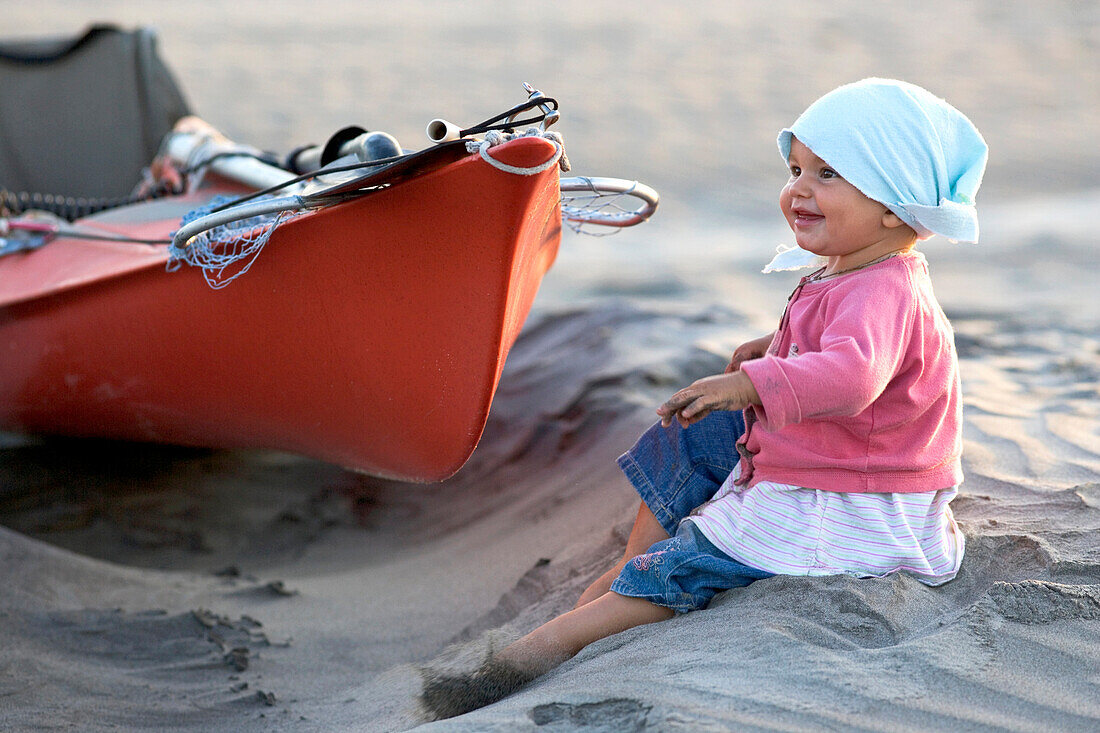Ein 1 Jähriges Mädchen sitzt am Strand bei einem Seakajak, Conejo, Baja Californien Süd, Mexiko