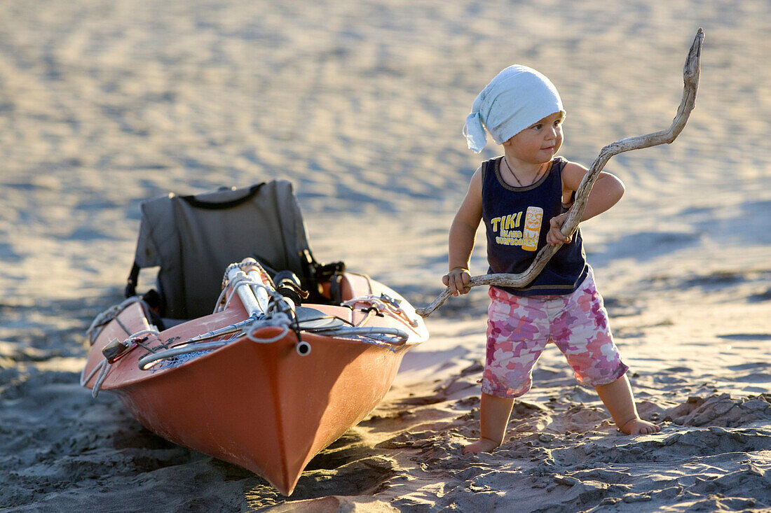 A 14 month old girl with a stick in her hand next to a sea kayak on the beach, Punta Conejo, Baja California Sur, Mexico