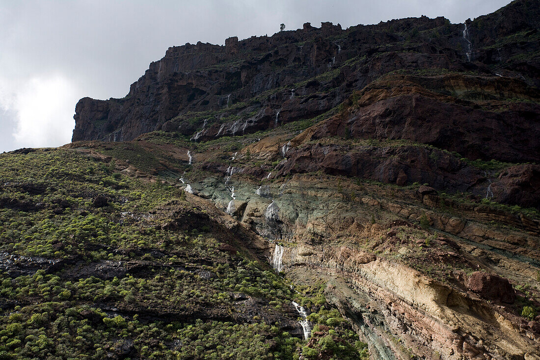 Waterfall and layers of volcanic rock under clouded sky, Los Azulejos, Barranco de Veneguera, Gran Canaria, Canary Islands, Spain, Europe